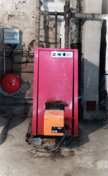 Chaudière au fioul ancienne de couleur rouge avec brûleur, installée dans une chaufferie avec des tuyaux de raccordement et un vase d'expansion.