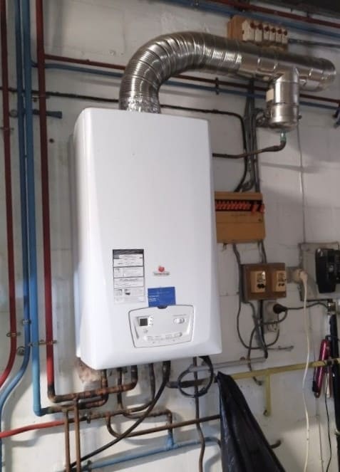 Chaudière murale à gaz avec conduit d’évacuation en inox, installée dans une salle technique avec raccordements hydrauliques et électriques.